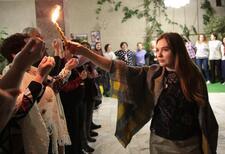 A young woman holding up a lit havdalah candle, walking around a room lined with other wome who hold up their fingernails to see the reflection of the light