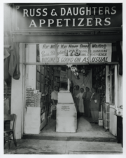 New York storefront, men standing inside. Sign reading: Russ & Daughters Appetizers. Second sign reading: For what have you been waiting...business going on as usual