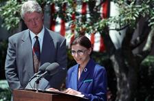 Ruth Bader Ginsburg speaking at a podium outdoors, with Bill Clinton standing behind her and an American flag in the background