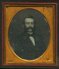 Photograph of a man in a circular frame, wearing a suit, with mutton chop-style sideburns and a moustache