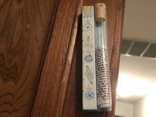 Mezuzah mounted on wooden door frame. Scroll visible, in a test tube-like vial corked on light blue wood decorated with pomegranates and Star of Davids.