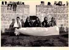 Four dancers sitting at a white cloth stretched between them in front of stone walls. Children sit on top of the walls.