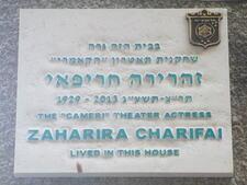 A white plaque with blue lettering in Hebrew and English. The English reads "1929-2013. The 'Cameri' Theater Actress Zaharira Charifai lived in this house."