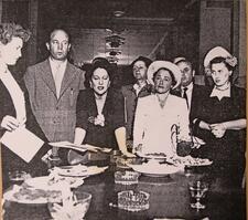 Bertha Kogan standing at a table, surrounded by seven other men and women