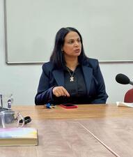 Woman with dark hair wearing dark suit sits at conference table; microphone point in her direction