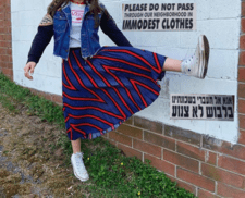 Young woman standing next to brick wall, wearing ankle-length blue skirt with red striped pattern and a Vans shirt underneath a jean jacket. Signs photoshopped into the image next to the young woman in English and Hebrew read: "Please do not pass through our neighborhood in immodest clothes" 