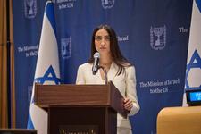 Woman with dark brown hair and white suit speaking into mic at podium, Israeli flag in background