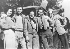 A group of two women and four men stand with arms around one another in front of a building.