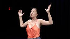 Woman in a theatrical performance, wearing orange tank stop and with arms in "hands up" position