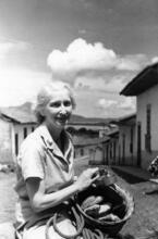 Doris Rosenthal sitting on a donkey in front of a residential street, holding a piece of equipment and a basket of bananas