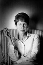 A portrait of anita Brookner, sitting in an armchair with her arms crossed, holding a cigarette