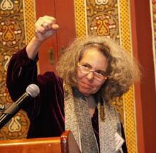 Writer and editor Melanie Kaye Kantrowitz standing at a podium making a clenched fist of solidarity