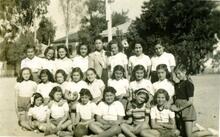 Three rows of girls wearing shorts and short-sleeved shirts, posing with their teacher in front of a building