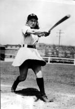 Thelma Eisen swinging a bat, wearing a skirted baseball uniform and a hat reading "GR"
