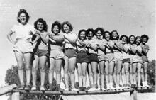 Thirteen young women stand, smiling and laughing, and pose with hands on each others' shoulders for a photo