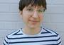 Young person with short brown hair and wire-rimmed glasses wearing a white t shirt with black stripes, posing against a white brick wall