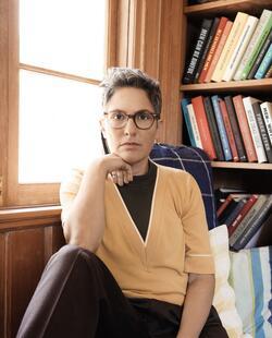 Joey Soloway wearing dark pants, yellow top, and glasses, seated in front of a window and a bookcase with right arm propped up on bent right leg