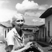 Doris Rosenthal sitting on a donkey in front of a residential street, holding a piece of equipment and a basket of bananas