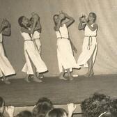 A group of six women, barefoot, wearing long white tunics, and holding up clay pots as they dance onstage