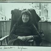 Denise Levertov sitting in a wooden chair, with potted flowers behind her