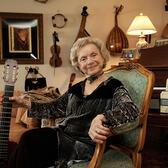 Flory Jagoda sitting in a chair, holding the neck of a guitar, with more instruments hanging on the wall behind her
