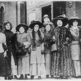 Nine women in long dresses, coats, and hats, with their arms linked, most smiling and laughing