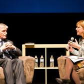 Heather Reisman sitting in an armchair, holding a microphone and speaking to a man seated across from her