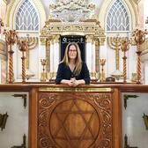 Mónica Unikel-Fasja standing before the Torah ark in an elaborately decorated synagogue with carved candelabras, menorahs, and columns.