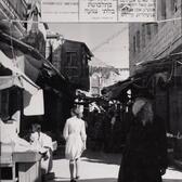 Photo of woman wearing a short dress in Mea Shearim, Israel, underneath a sign mandating modest dress. A man covers his eyes as she passes.