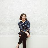 Niki Russ Federman smiling, seated with legs crossed on a white bench in front of a white textured wall