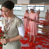 A dancer in a patterned dress holding a rose in a dance studio with a mirrored wall. Behind her stand four other dancers in pink dresses and white headscarves, holding red rose petals, which are scattered heavily over the floor.