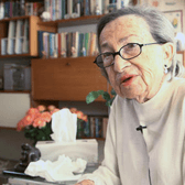 Vele Zabludovsky sitting in a living room, with a microphone clipped to her sweater