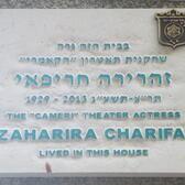 A white plaque with blue lettering in Hebrew and English. The English reads "1929-2013. The 'Cameri' Theater Actress Zaharira Charifai lived in this house."