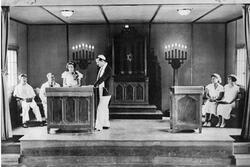 A picture of a bimah, with two men and two women sitting on either side, and a man in a kippah and prayer shawl standing by a podium, and a young woman with flowers pinned to her dress speaking at the podium