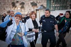Two women wearing tallit are moving away from the wall, a police officer looking at them and speaking to them. Another woman films the event on her phone.