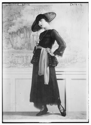 Fannie Brice standing wearing a large dark colored dress and large hat, hand on her long necklace