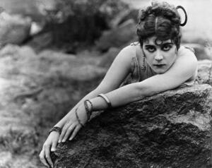 Theda Bara in makeup and costume with many bracelets, posed on a large rock with her head resting on her left arm