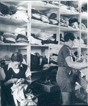 Two women, one sitting and mending a piece and one standing, in the kibbutz clothing warehouse, in front of a wall of shelves with stacks of clothing. 