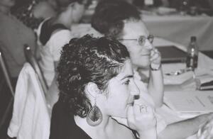 Profile photo with focus on a woman with her hand on her chin. A man seated beside her slightly out of focus in background.