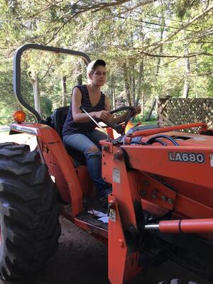 Janna Siller wearing ripped jeans, tank top, and baseball cap, driving an orange tractor