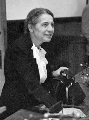 Lise Meitner standing at a table beside an atomic model, holding a purse and briefcase in one hand