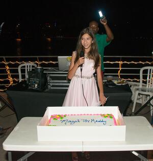 Maddy Pollack speaking at her Bat Mitzvah party, microphone in hand. Cake on a table in front of her with text "Mazel Tov, Maddy."