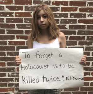High school student standing in front of a brick wall. She is holding a protest sign that says "To forget a Holocaust is to be killed twice," attributed to Elie Wiesel.