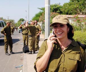 Miri Regev, in soldier's uniform, smiling while talking on the phone on the side of the road, with other soldiers standing around in background.