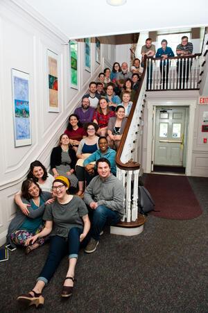 A group of about twenty-five people sitting on a staircase, with some leaning against the rail on the second floor landing