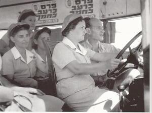 A woman sits at the wheel of a bus with a man next to her instructing, with three women behind them observing.
