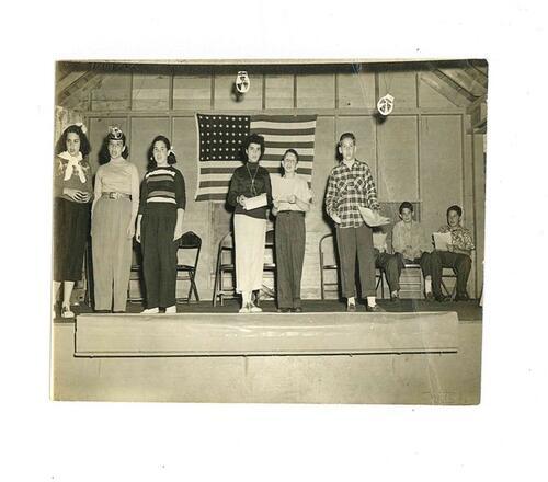 Children on stage in front of an American flag for play rehearsal at Berkshire Hills Camp. Joan Mykoff, center stage in a group of six. 