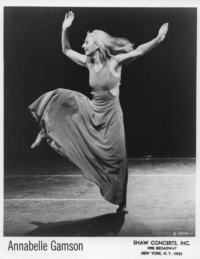 Annabelle Gamson, on stage, wearing a long dress and lifting both arms and one leg in the air