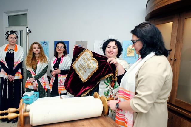 Woman removes cover from Torah scroll at bimah as group of women looks on