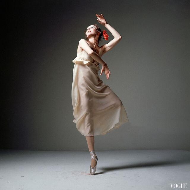 A photo of Allegra Kent standing on one foot en pointe in an empty studio, wearing a gauzy cream-colored dress with two red flowers in her hair